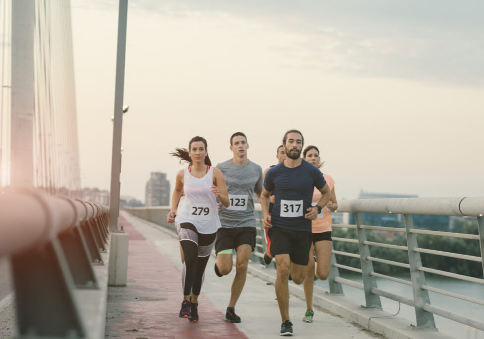 Runners running marathon in the city. They are running over the bridge at sunset. Wearing numbers on their sport clothes. Cityscape in background.