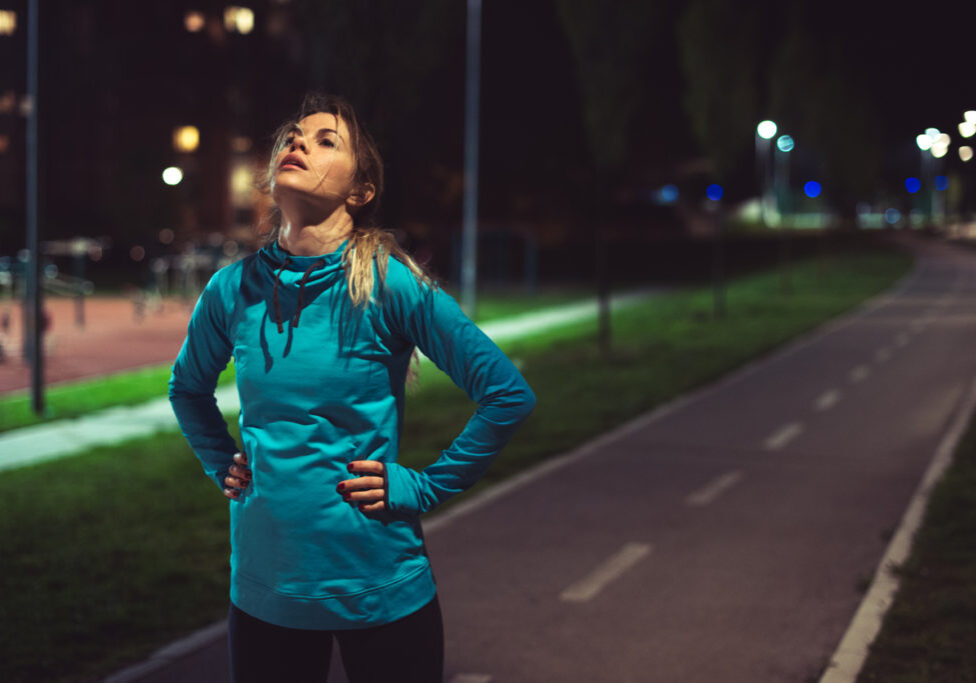 Young tired woman standing on the running track outdoor after running. It's night time.