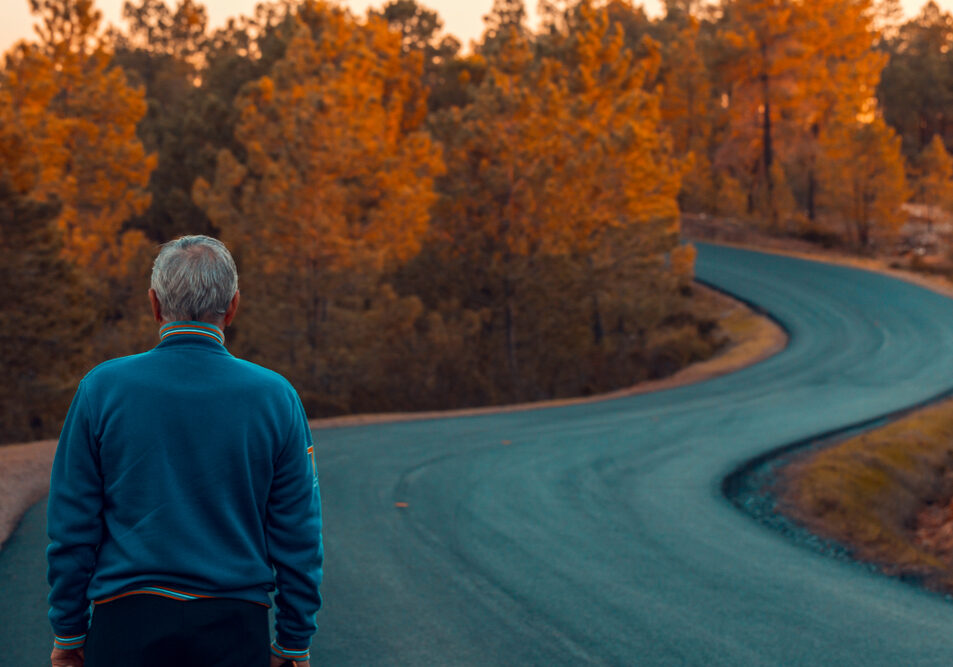 Active senior man stands alone on lonely road between mountains. Older man of back walking on lonely highway