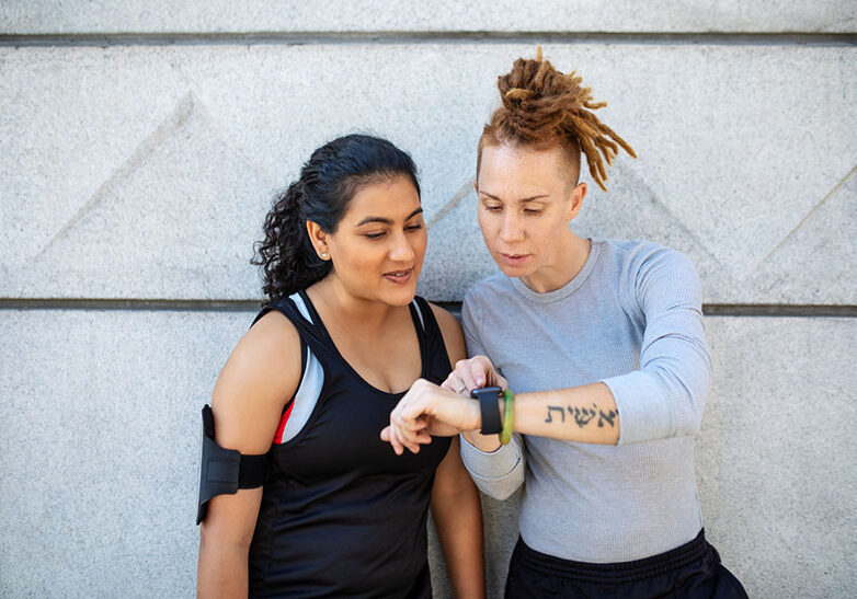 Two sporty women wearing sportswear working out outdoors in the city having a break and looking at smartwatch. Female friends looking at fitness tracker.