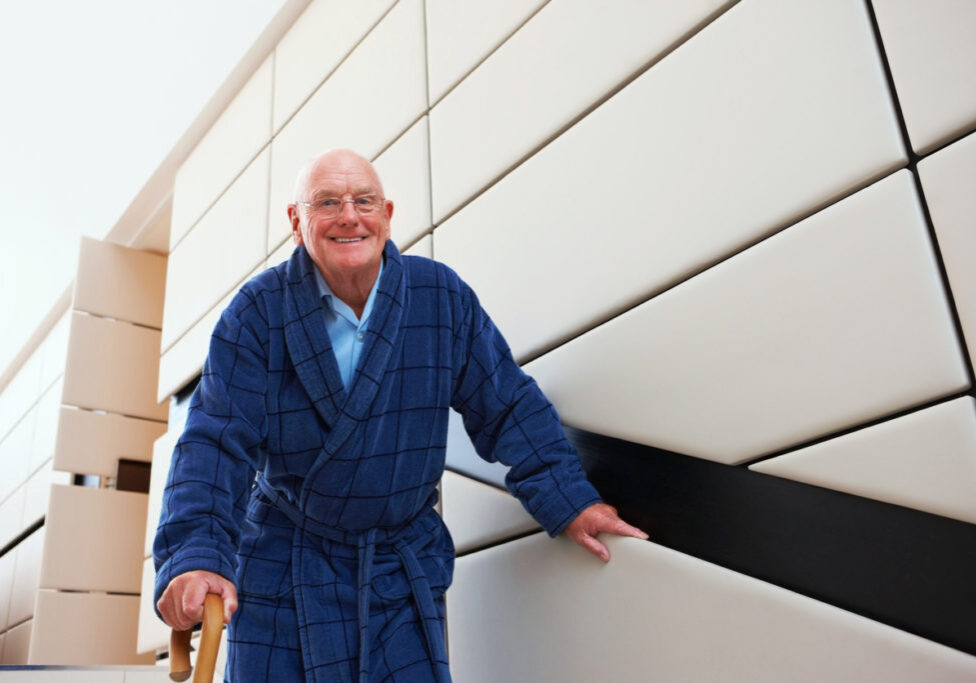 Elderly man in bathrobe walking down the stairs with help of walking stick looking at camera smiling