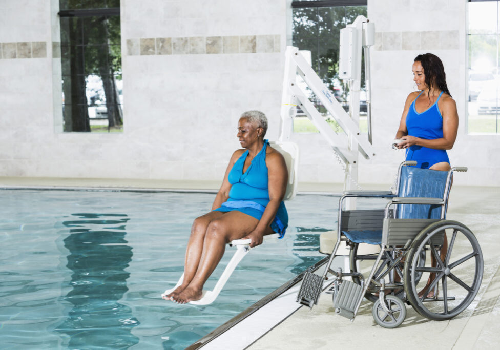 A patient undergoing hydrotherapy treatment in a swimming pool.  A physical therapist is lowering a senior African American woman into the water on a special lift.  The patient's wheelchair is on the pool deck.
